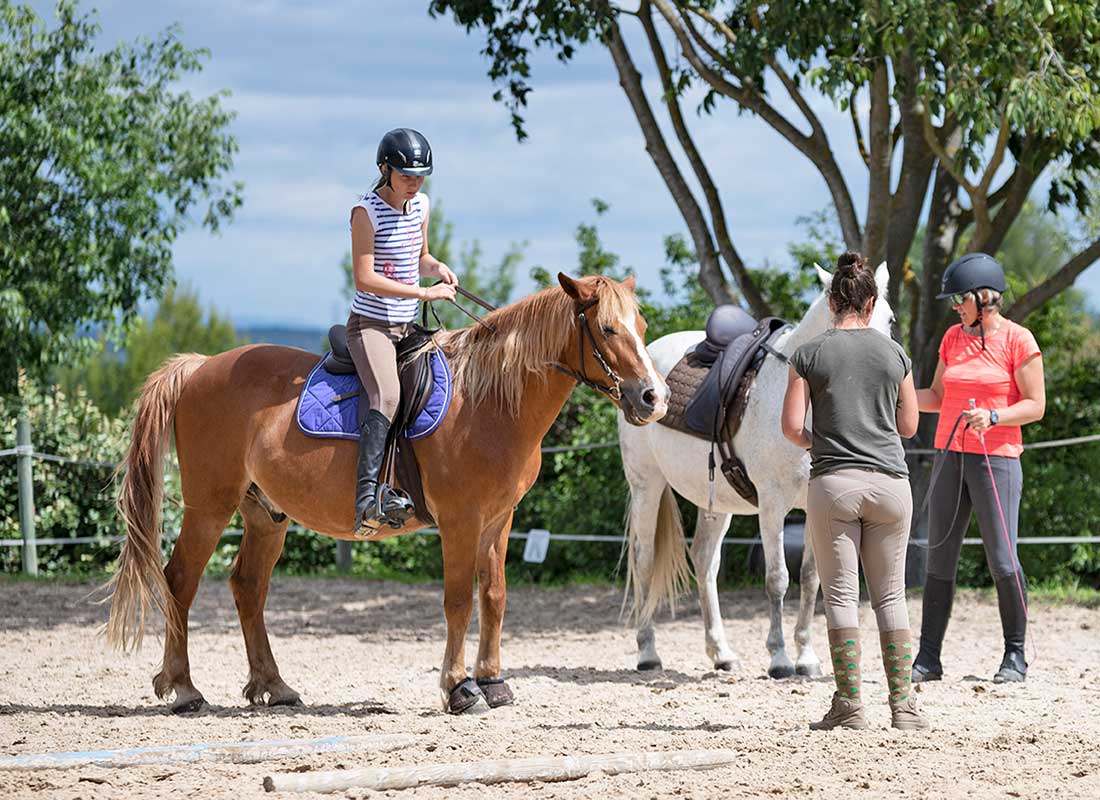 Girl riding a brown horse with saddle, two women standing nearby, outdoor riding lesson setting, surrounded by trees and blue sky, highlighting equestrian activities near Liberty Farm's attractions.